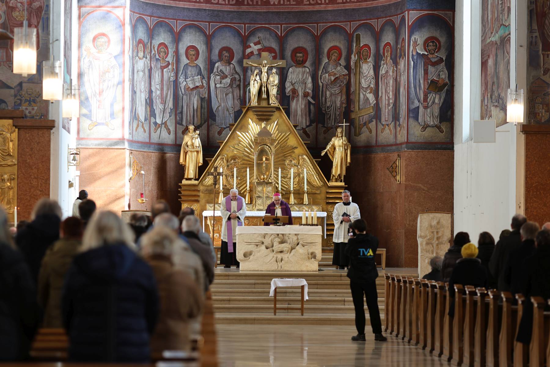 Bischof Rudolf Voderholzer steht mit zwei Geistlichen vor dem Altar in der Kirche St. Anton, umgeben von Wandmalereien mit Heiligenfiguren und einer goldenen Altarverkleidung.